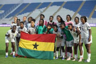 Ghana women's soccer team posing on the field with a Ghana flag in front of them on a stadium pitch.