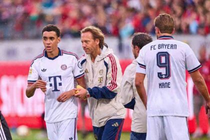 Bayern Munich midfielder Jamal Musiala wearing a white away kit is assisted off the pitch by FC Bayern team doctor Prof. Dr. Peter Ueblacker after falling during a preseason friendly between 1. FC Heidenheim and FC Bayern Munich at Voith-Arena on August 18, 2026 in Heidenheim, Germany. Harry Kane #9 is seen in the background