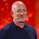 Bald man wearing a navy polo on stage with a red glittery background and a small headset microphone on his cheek  trybalnews