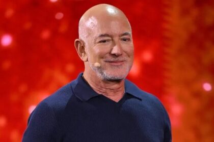 Bald man wearing a navy polo on stage with a red glittery background and a small headset microphone on his cheek.