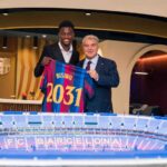 FC Barcelona player Jesse Bisiwu and Barcelona president Joan Laporta pose indoors holding up a blue and red Barcelona jersey with BISIWU and 2031 printed on the back after Bisiwu signed a six year contract with the club A model of the FC Barcelona stadium sits on the table in front of them and the Barcelona crest is on the wall behind  trybalnews