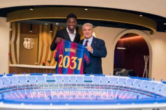 FC Barcelona player Jesse Bisiwu and Barcelona president Joan Laporta pose indoors holding up a blue and red Barcelona jersey with "BISIWU" and "2031" printed on the back, after Bisiwu signed a six year contract with the club. A model of the FC Barcelona stadium sits on the table in front of them and the Barcelona crest is on the wall behind.