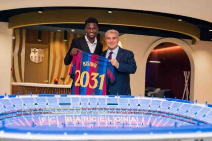 FC Barcelona player Jesse Bisiwu and Barcelona president Joan Laporta pose indoors holding up a blue and red Barcelona jersey with "BISIWU" and "2031" printed on the back, after Bisiwu signed a six year contract with the club. A model of the FC Barcelona stadium sits on the table in front of them and the Barcelona crest is on the wall behind.