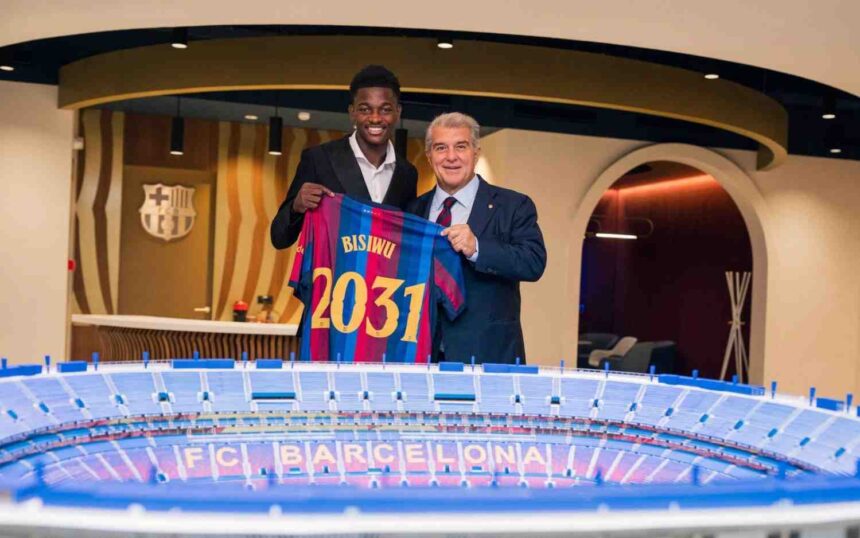 FC Barcelona player Jesse Bisiwu and Barcelona president Joan Laporta pose indoors holding up a blue and red Barcelona jersey with BISIWU and 2031 printed on the back after Bisiwu signed a six year contract with the club A model of the FC Barcelona stadium sits on the table in front of them and the Barcelona crest is on the wall behind  trybalnews