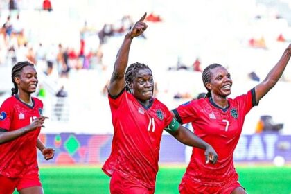 Three female soccer players in red jerseys celebrate on a green pitch, arms raised in triumph, with a blurred stadium crowd behind them.