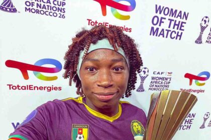 Female Cameroon footballer selfies after a win, wearing a purple jersey and headband, holding a tall gold trophy.