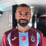 Man with short dark hair and beard posing for a selfie in an airplane cabin wearing a maroon and light blue football jersey with a Turkish flag emblem and a team crest on the chest  trybalnews