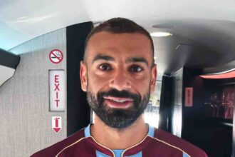Man with short dark hair and beard posing for a selfie in an airplane cabin wearing a maroon and light blue football jersey with a Turkish flag emblem and a team crest on the chest  trybalnews