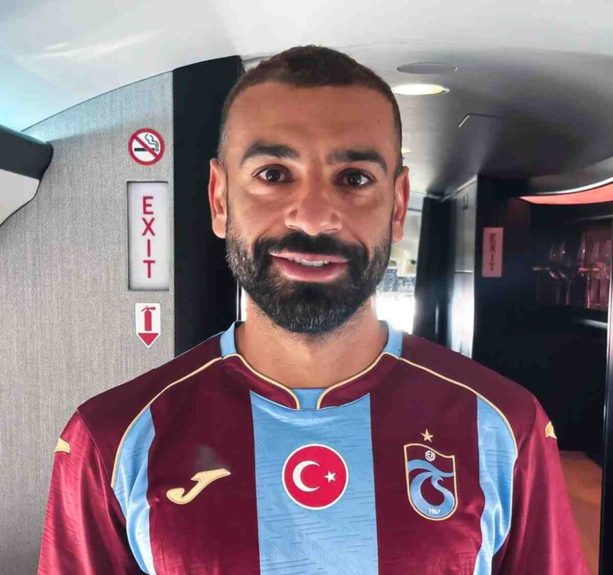 Man with short dark hair and beard posing for a selfie in an airplane cabin, wearing a maroon and light-blue football jersey with a Turkish flag emblem and a team crest on the chest.