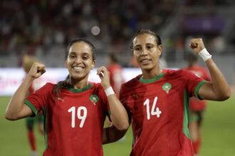 Two female soccer players in red jerseys celebrate on the field, fists raised in victory.