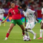 Female soccer players compete for the ball during a match player in red jersey number 2 and green shorts challenges a white jersey opponent near the ball on a green field  trybalnews