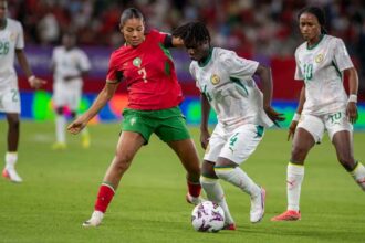Female soccer players compete for the ball during a match; player in red jersey number 2 and green shorts challenges a white-jersey opponent near the ball on a green field.