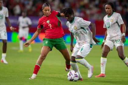 Female soccer players compete for the ball during a match; player in red jersey number 2 and green shorts challenges a white-jersey opponent near the ball on a green field.