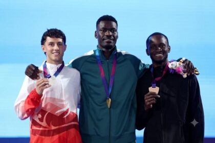 Gold medalist Ezekiel Nathaniel of Team Nigeria in green tracksuit stands on the podium with silver medalist Jake Minshull of Team England in red and white jacket on the left and bronze medalist Assinie Wilson of Team Jamaica in black tracksuit on the right during the Men's 400 Metres Hurdles Final medal ceremony on Day 8 of the Glasgow 2026 Commonwealth Games at Scotstoun Stadium, July 31, 2026.