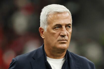 Older man with silver hair wearing a dark blazer and white shirt, looking to the side, with a blurred stadium crowd in the background.