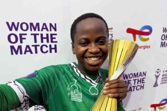 Smiling female athlete in a green jersey holds a gold trophy at a 'Woman of the Match' event backdrop.