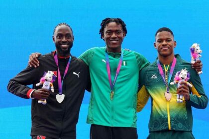 Gold medalist Samuel Ogazi of Team Nigeria in a green Puma tracksuit with the Nigerian flag stands in the center on the podium with his arm around silver medalist Jereem Richards of Trinidad and Tobago in a black jacket on the left and bronze medalist Zakithi Nene of South Africa in a green and yellow tracksuit on the right. All three hold medals and Commonwealth Games mascots during the Men's 400 Metres Final medal ceremony on Day 9 of the Glasgow 2026 Commonwealth Games at Scotstoun Stadium, August 1, 2026.