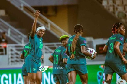 Nigeria's Super Falcons player Monday Gift, wearing jersey number 20 and a white headband, raises her hand and celebrates with teammates after scoring the team's second goal during a WAFCON Group C match against Egypt. Other Nigerian players in teal and green kits are running and celebrating on the pitch in a stadium.