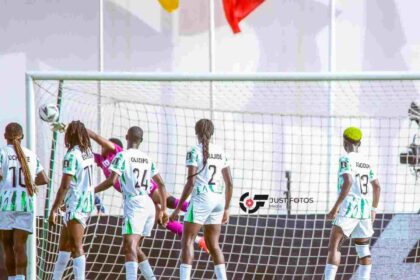 Women’s soccer team in white and green uniforms gathers near the goal, celebrating a moment near the net as the goalkeeper wears pink.