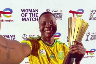 Female soccer player in a yellow jersey posing with a tall gold trophy, smiling, against a 'Woman of the Match' backdrop.