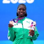 Female athlete standing on a podium holding a star shaped medal and a mascot trophy wearing a green jacket and smiling against a blue background at the event  trybalnews