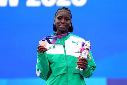 Female athlete standing on a podium, holding a star-shaped medal and a mascot trophy, wearing a green jacket and smiling against a blue background at the event.