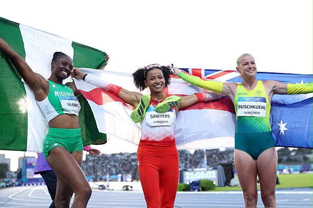 Glasgow 2026 Trybal News Three female athletes stand on a track at the finish area arms linked in celebration with large national flags visible behind them trybalnews
