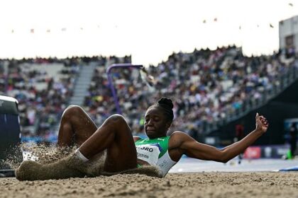 Female track and field athlete lands in the sand pit during a long jump, arms extended, with a crowded stadium in the background.