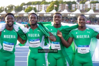 Four Nigerian male sprinters in green Nigeria jerseys pose with arms around each other after a race, smiling on a track with crowd in the background.