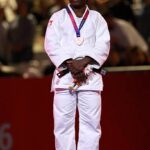 Female judoka in a white gi stands on a podium with a silver medal hanging from a purple ribbon spectators blurred in the background  trybalnews