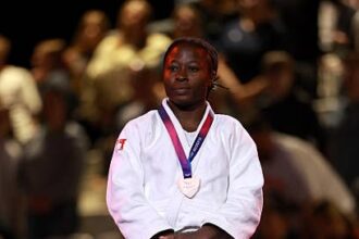 Female judoka in a white gi stands on a podium with a silver medal hanging from a purple ribbon, spectators blurred in the background.