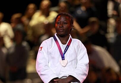 Female judoka in a white gi stands on a podium with a silver medal hanging from a purple ribbon, spectators blurred in the background.