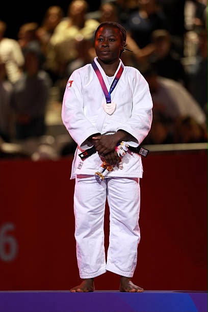 Female judoka in a white gi stands on a podium with a silver medal hanging from a purple ribbon spectators blurred in the background  trybalnews
