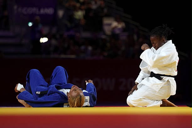 Glasgow 2026 Trybal News Judo match athlete in blue lies on her back on the mat while the opponent in white kneels beside her trybalnews