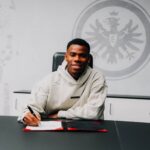 Smiling man in a beige hoodie signs papers at a desk with a large emblem on the wall behind him  trybalnews