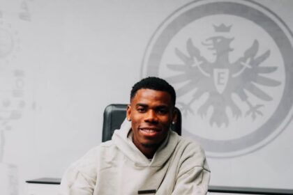 Smiling man in a beige hoodie signs papers at a desk, with a large emblem on the wall behind him.