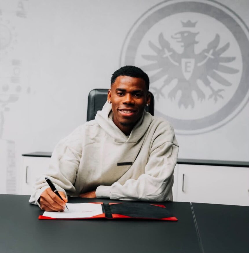 Smiling man in a beige hoodie signs papers at a desk with a large emblem on the wall behind him  trybalnews