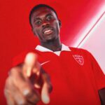 Young man in a red polo shirt points toward the camera smiling against a red wall backdrop  trybalnews