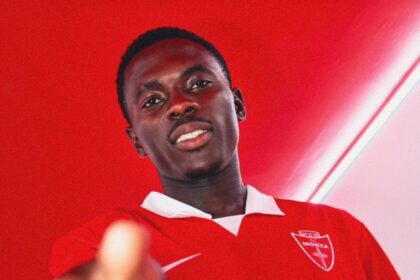Young man in a red polo shirt points toward the camera, smiling, against a red wall backdrop.