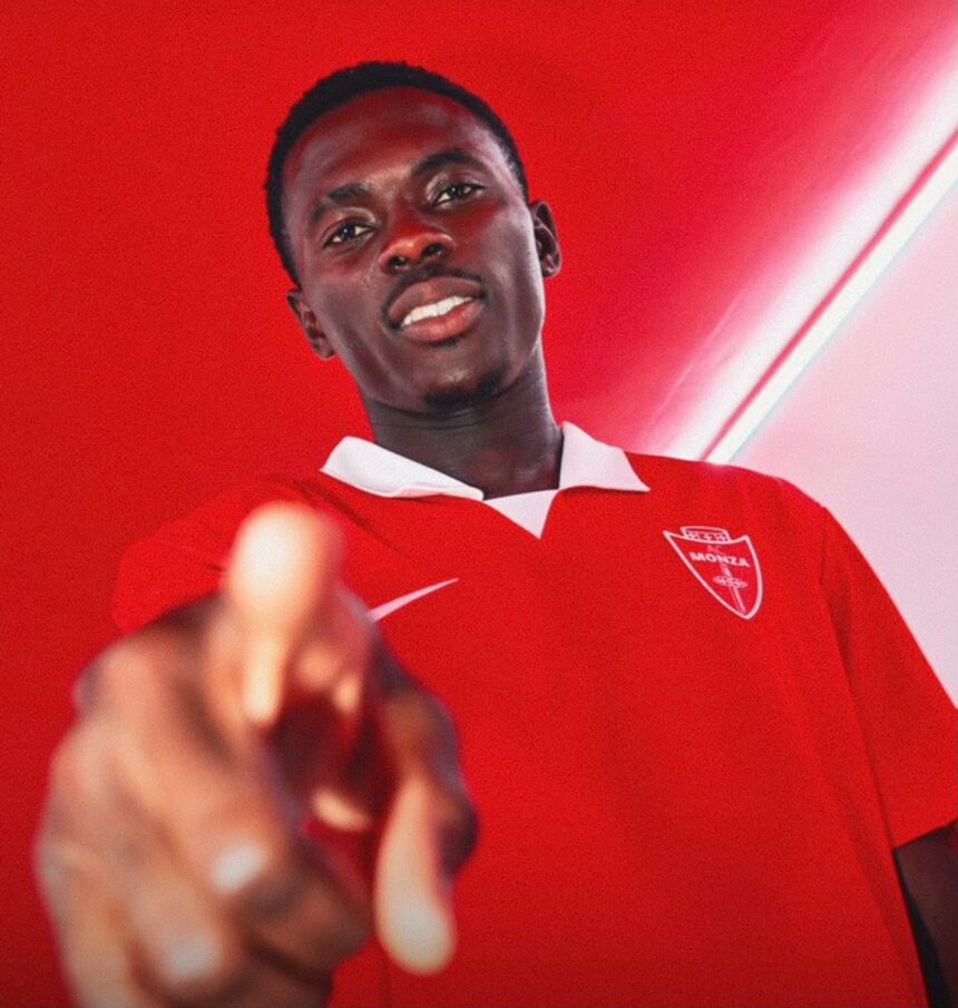 Young man in a red polo shirt points toward the camera smiling against a red wall backdrop  trybalnews