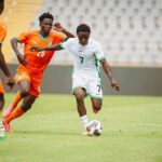 Soccer players in orange and white chase a ball on a green field during a CAF U 20 Africa Cup qualifying match with one player in white dribbling  trybalnews
