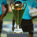 Gold trophy with two handles on a pedestal labeled CAP Conference Cup 2026 foreground with blurred players in teal shirts in the background  trybalnews
