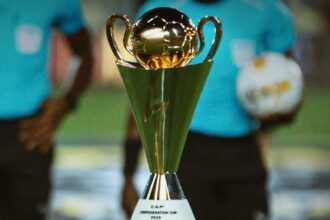 Gold trophy with two handles on a pedestal, labeled 'CAP Conference Cup 2026', foreground with blurred players in teal shirts in the background.