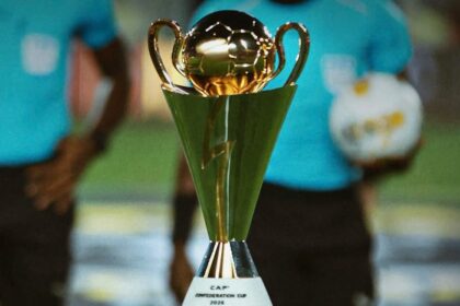 Gold trophy with two handles on a pedestal, labeled 'CAP Conference Cup 2026', foreground with blurred players in teal shirts in the background.