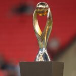 Golden trophy with a spherical top and curved arms on a square base against a red blurred background  trybalnews