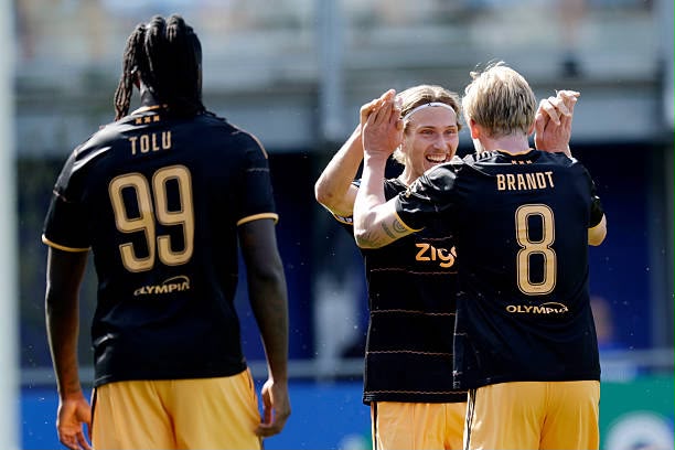 Three soccer players in black and gold uniforms celebrate on the field one with hands raised and another embracing with jersey numbers 99 Tolu and 8 Brandt visible on their backs  trybalnews