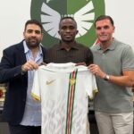 Three men pose holding a white soccer jersey with a gold Nike swoosh and a greenredgreen vertical stripe in front of a club crest  trybalnews