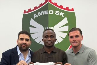 Three men pose holding a white soccer jersey with a gold Nike swoosh and a green–red–green vertical stripe in front of a club crest.
