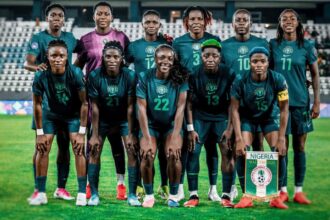 Nigeria women's national football team group photo on the pitch in teal kits, with a Nigeria banner visible in front.