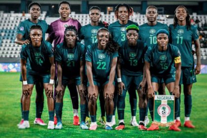 Nigeria women's national football team group photo on the pitch in teal kits, with a Nigeria banner visible in front.